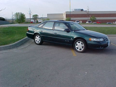 1994 Ford Taurus - A dark green 1994 Ford Taurus is parked in an asphalt lot with a building and trees in the background.