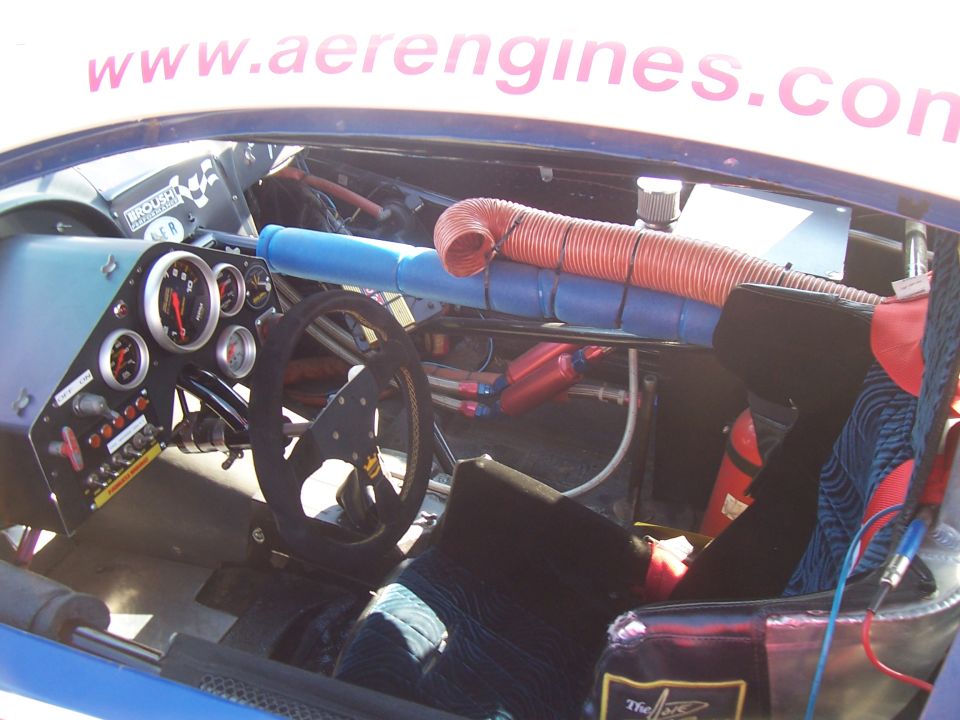 Racing car interior with custom dashboard, steering wheel, and roll cage. Red and orange hoses are visible