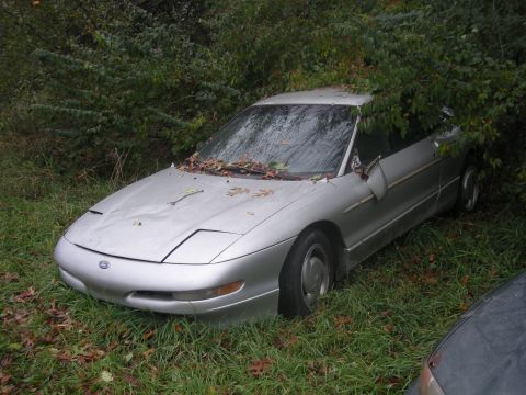 1996 Ford Probe - Abandoned silver coupe with pop-up headlights sits in overgrown grass and bushes.