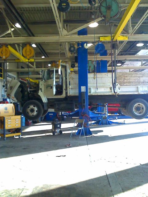 1992 Ford Bronco - A white flatbed truck is suspended on a lift inside a garage, with its hood open for repairs.