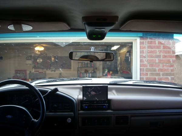 Interior view of a vehicle dashboard with a steering wheel, aftermarket stereo, and a rearview mirror. Garage visible through windshield