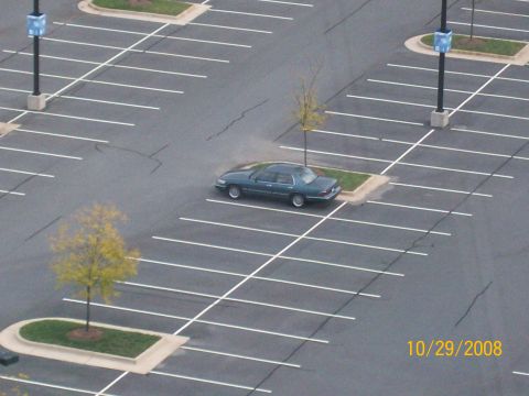 1995 Mercury Grand Marquis - A lone sedan sits in a vast, empty parking lot, marked by white lines and a few small trees.