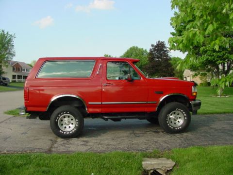 1992 Ford Bronco - A red 1992 Ford Bronco is parked on a paved surface with grass and trees in the background.