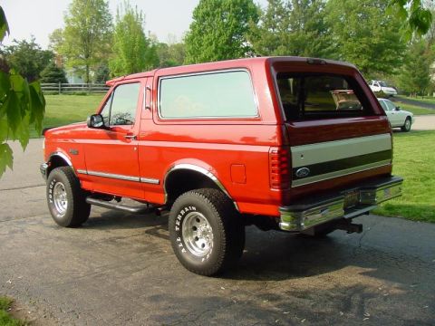 1992 Ford Bronco - This red 1992 Ford Bronco is shown from a rear three-quarter view, parked on a paved surface with a grassy area and trees in the background.