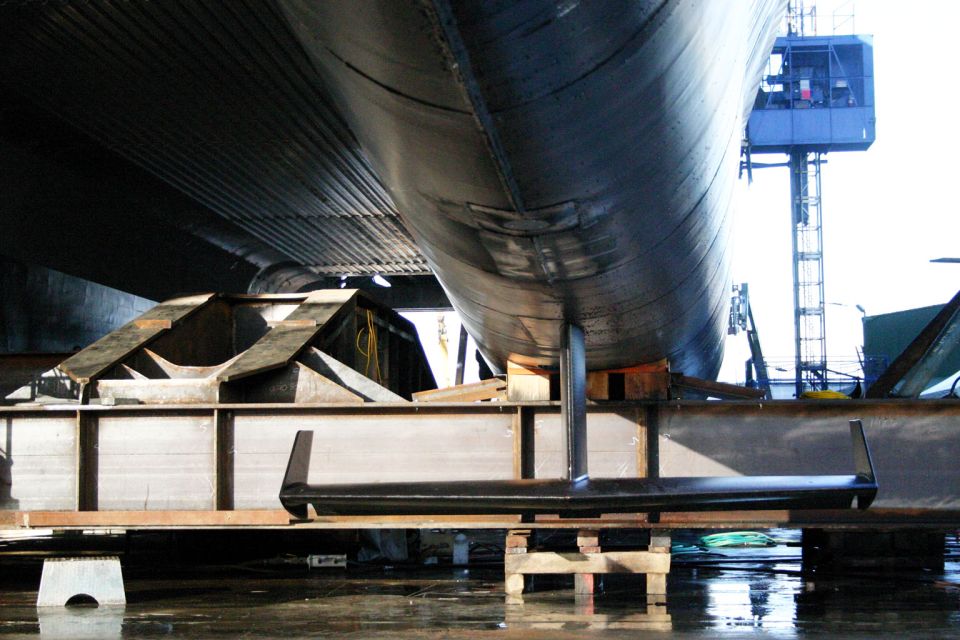 Underside of a large hull on supports, with a metal structure and a crane in the background