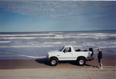 1992 Ford Bronco - A white 1992 Ford Bronco is parked on the sand near the ocean waves.