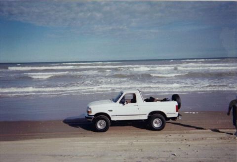 1992 Ford Bronco - A white 1992 Ford Bronco sits on the sand near the ocean waves under a partly cloudy sky.