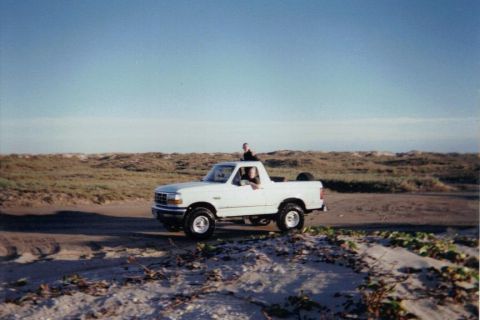 1992 Ford Bronco - A white 1992 Ford Bronco drives along a sandy path with people in and on the vehicle, set against a backdrop of dunes and a clear sky.