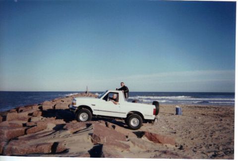 1992 Ford Bronco - A white 1992 Ford Bronco is parked on a rocky shoreline with the ocean in the background.