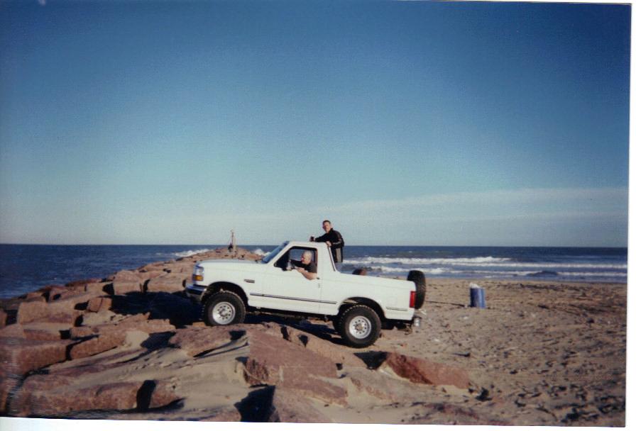 White 1992 Ford Bronco parked on rocks by the ocean, with two people visible