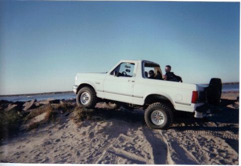 1992 Ford Bronco - A white 1992 Ford Bronco navigates a sandy incline near the coast on a clear day.