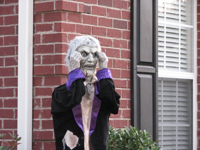 Scary Halloween decoration of a zombie figure with white hair and glowing eyes, standing in front of a brick house