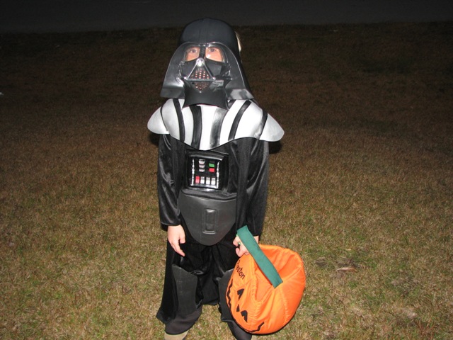 Child dressed as Darth Vader for Halloween, holding an orange pumpkin candy bucket