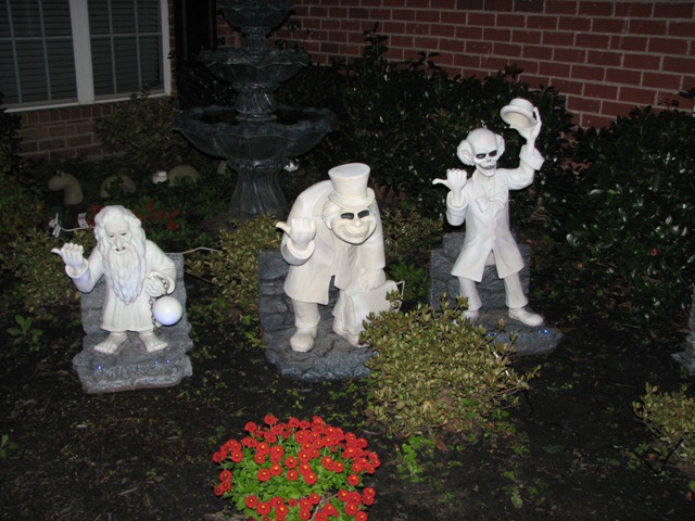 Three white haunted mansion character statues in a yard with a fountain and red flowers