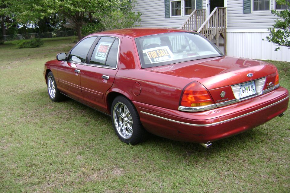 Red Ford Crown Victoria sedan on grass, rear three-quarter view, chrome wheels and For Sale signs in windows