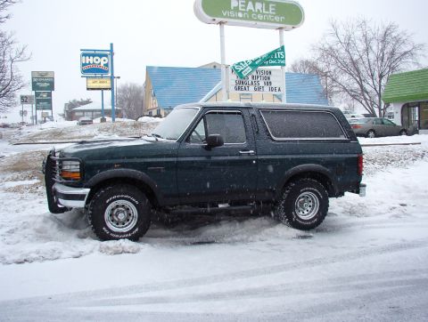 1993 Ford Bronco - This dark green SUV is parked in a snowy lot, with businesses visible behind it.