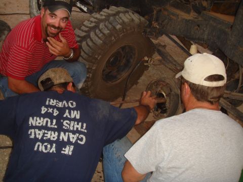 1992 Ford Bronco - Three men are gathered around the muddy undercarriage and tire of a 4x4 vehicle, seemingly in the process of repair.