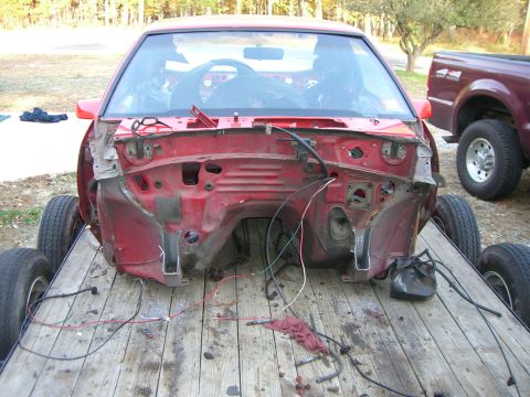 1988 Ford Bronco - The front end of a red car, missing its engine and body panels, sits on a trailer. A truck is parked nearby.