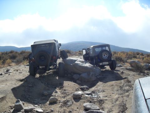 1990 Ford Bronco - Two Jeeps tackle a challenging off-road course with rocky terrain and steep inclines under a partly cloudy sky.