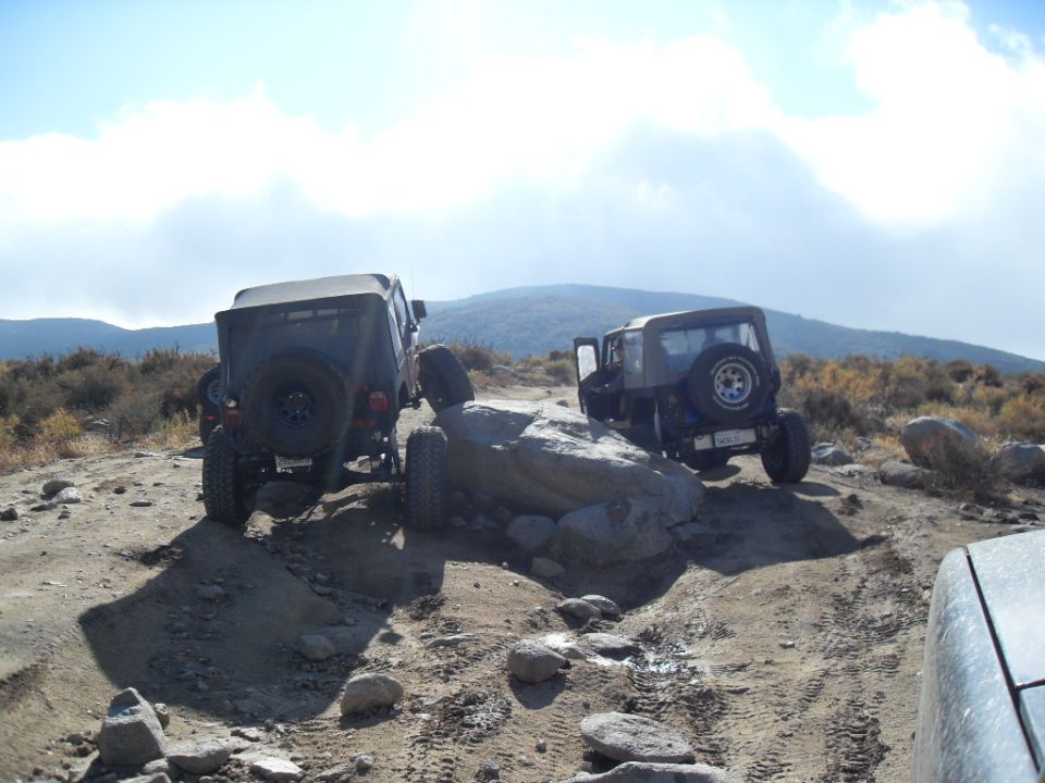 Two Jeeps are navigating a rocky, off-road trail. One is on the left, the other is on the right, partially behind a large rock