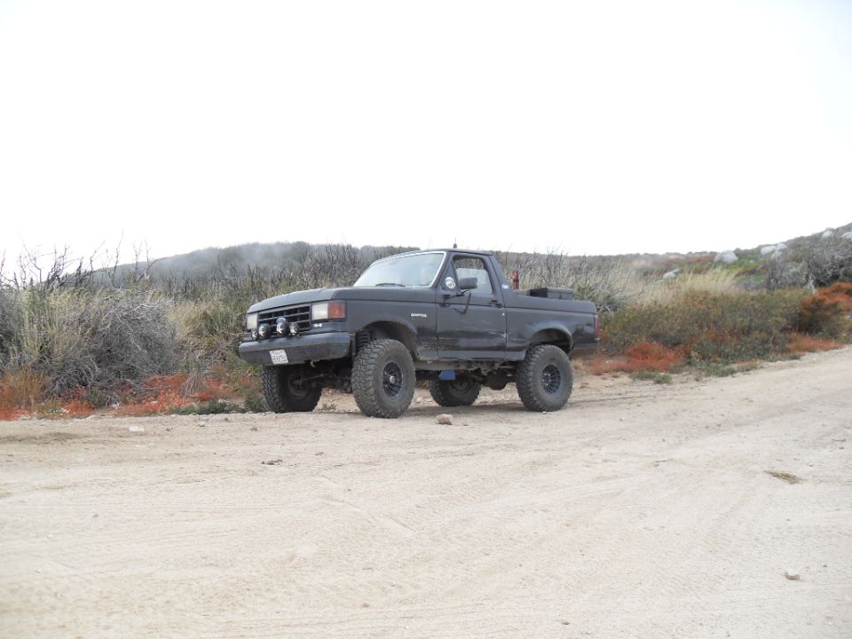 Black lifted truck parked on a dirt road, front-left quarter view. It has aftermarket lights and large tires