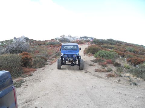 1990 Ford Bronco - A blue Jeep Wrangler navigates a dirt path in a rocky, arid landscape, with another vehicle partially visible on the left.