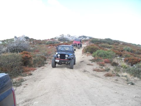 1990 Ford Bronco - Jeeps navigate a dusty trail through a rocky, scrub-covered hillside under a bright sky.