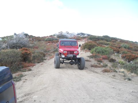 1990 Ford Bronco - A red Jeep Wrangler drives up a dusty trail in a rocky, arid landscape.