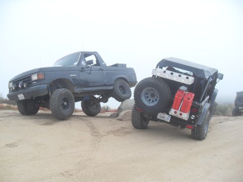 1990 Ford Bronco - Two modified off-road vehicles are positioned on a sandy, rocky area with a foggy background.