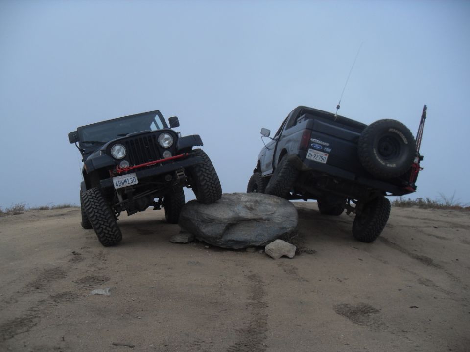 Black 1990 Ford Bronco with spare tire on rear, angled on a rock, with a black Jeep in front
