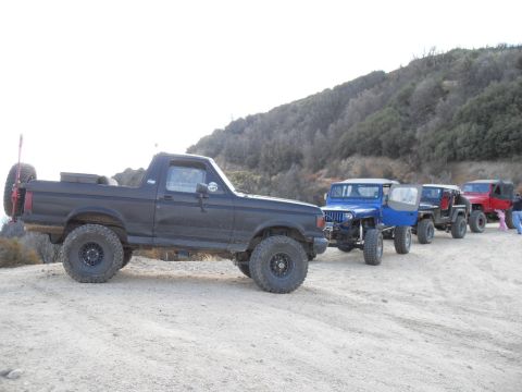 1990 Ford Bronco - A line of off-road vehicles, including a 1990 Ford Bronco, parked on a dirt trail.
