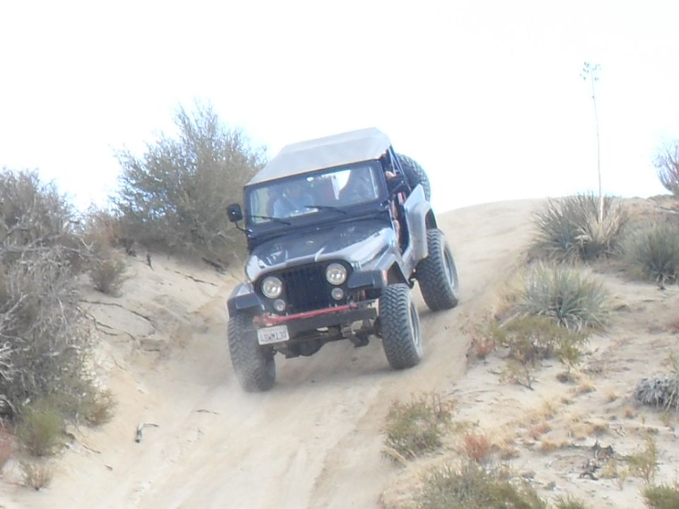 Black Jeep CJ-7 with a tan soft top driving up a sandy incline, red front bumper, California license plate