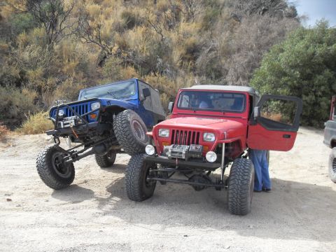 1990 Ford Bronco - Two modified Jeep Wranglers are parked off-road, one red and one blue, with the red Jeep's door ajar.