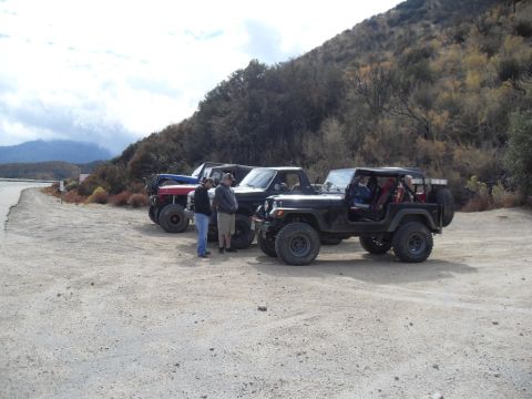 1990 Ford Bronco - Several modified off-road vehicles are parked together on a dusty lot near a hillside.