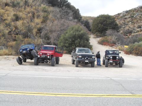 1990 Ford Bronco - A group of four modified off-road vehicles, including a blue Jeep, a red Jeep, a black pickup truck, and a black Jeep, are parked on a dirt…