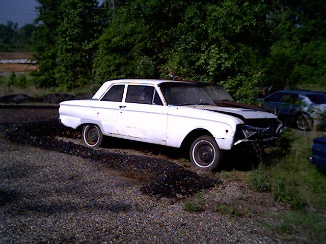 White vintage Ford Falcon coupe with front-end crash damage on a gravel lot.