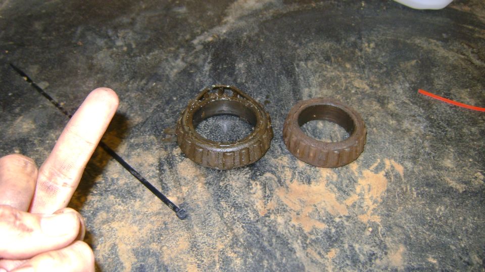 Two used wheel bearings, one with cage damage, on a dirty surface. A finger points to the left