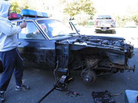 2001 Ford Crown Victoria - A dark sedan, possibly a former police vehicle, has its front end disassembled while a person works on it, with a pickup truck visible in t…