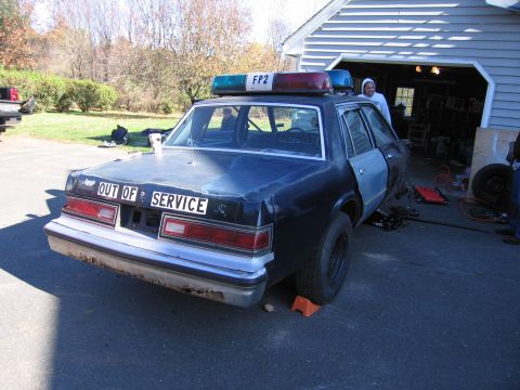 2001 Ford Crown Victoria - A dark-colored sedan, possibly a former police vehicle, is parked outside a garage with its rear facing the camera.