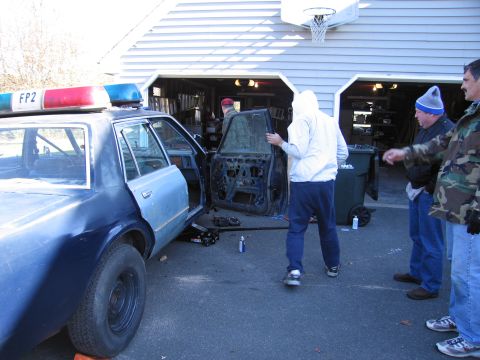 2001 Ford Crown Victoria - A police car, possibly a Ford Crown Victoria, is seen with its doors removed as people work on it in a driveway.