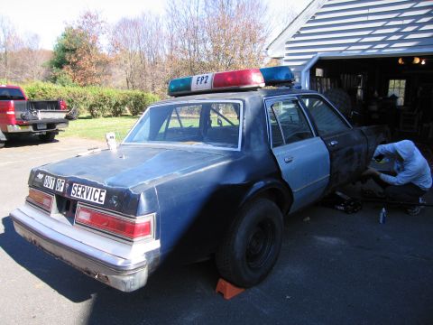 2001 Ford Crown Victoria - A police car is undergoing repairs in a driveway, with a red pickup truck visible in the background.