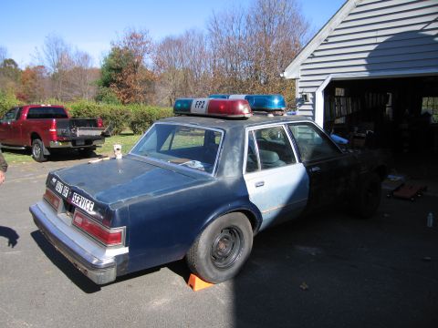 2001 Ford Crown Victoria - A weathered police car with a light bar sits in a driveway next to a red pickup truck.
