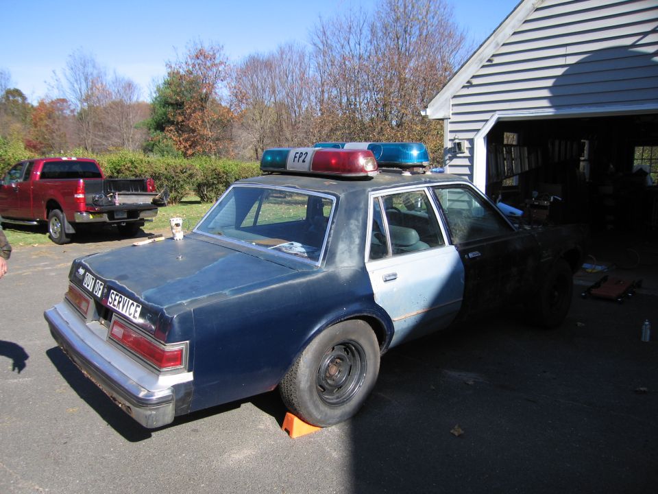 Old police car with light bar and 'OUT OF SERVICE' text on the trunk. Red pickup truck in background