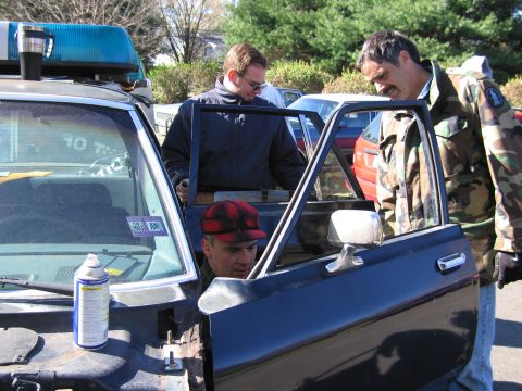 2001 Ford Crown Victoria - Three men are gathered around the open door of a police car, with a spray can visible on the hood.