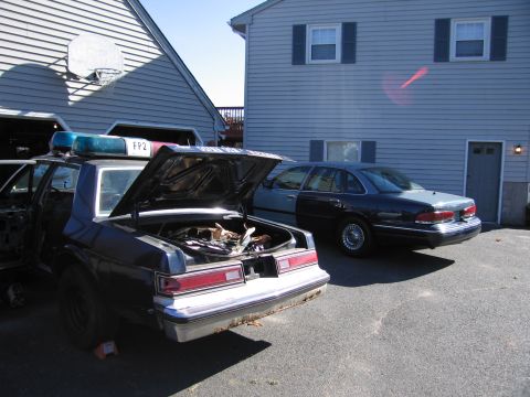 2001 Ford Crown Victoria - Two sedans, one a police car with an open trunk, are parked in a driveway.
