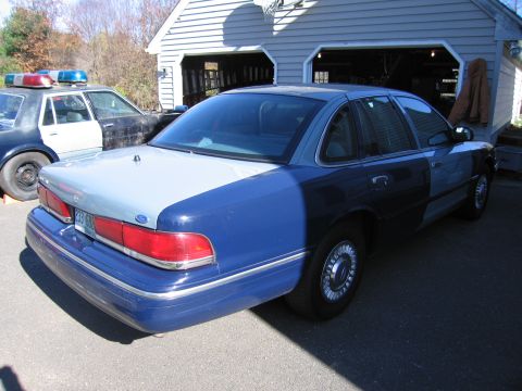 2001 Ford Crown Victoria - A two-tone Ford Crown Victoria police car is parked next to another police vehicle in front of a garage.