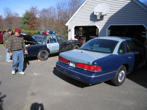 2001 Ford Crown Victoria - Two police cars are parked outside a garage, one with its trunk open.