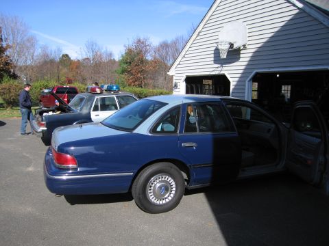 2001 Ford Crown Victoria - A blue and light blue 2001 Ford Crown Victoria sedan is parked in front of a garage with its doors open. Another police car is visible in t…