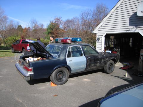 2001 Ford Crown Victoria - Side view of a police car with its trunk open, parked outside a garage with a red truck nearby.
