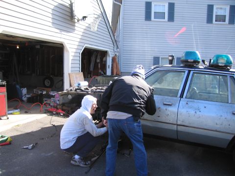 2001 Ford Crown Victoria - Two individuals are working on a weathered vehicle, possibly a former police car, in a driveway.